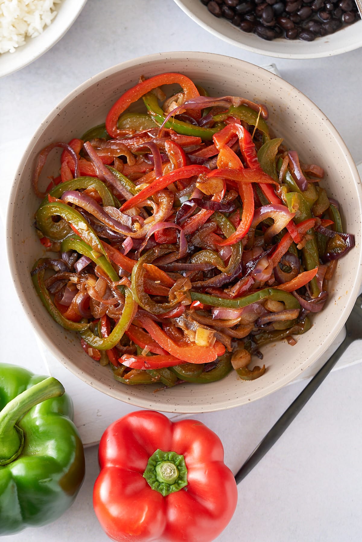 A large serving dish filled with fajitas vegetables, with a red and green bell peppers, and bowls of steamed white rice and black beans set alongside.