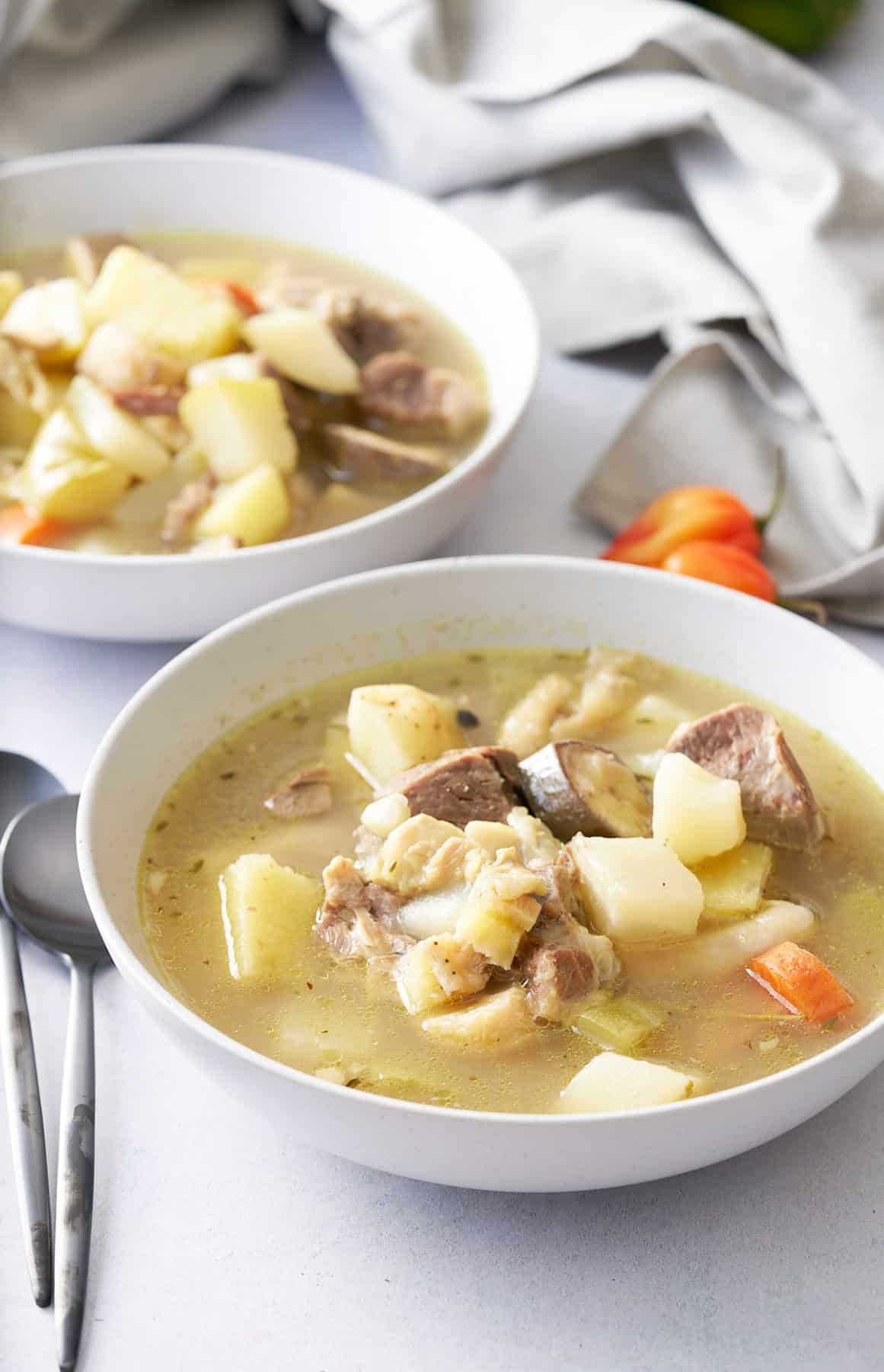 Two white bowls filled with goat, tripe and vegetable soup, with spoons, and Scotch bonnet peppers set alongside.