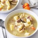 Two white bowls filled with goat, tripe and vegetable soup, with spoons, and Scotch bonnet peppers set alongside.