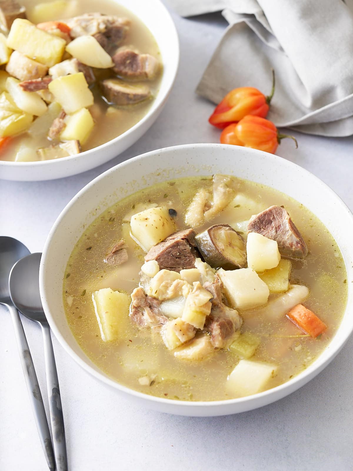 Two white bowls filled with goat, tripe and vegetable soup, with spoons, and Scotch bonnet peppers set alongside.