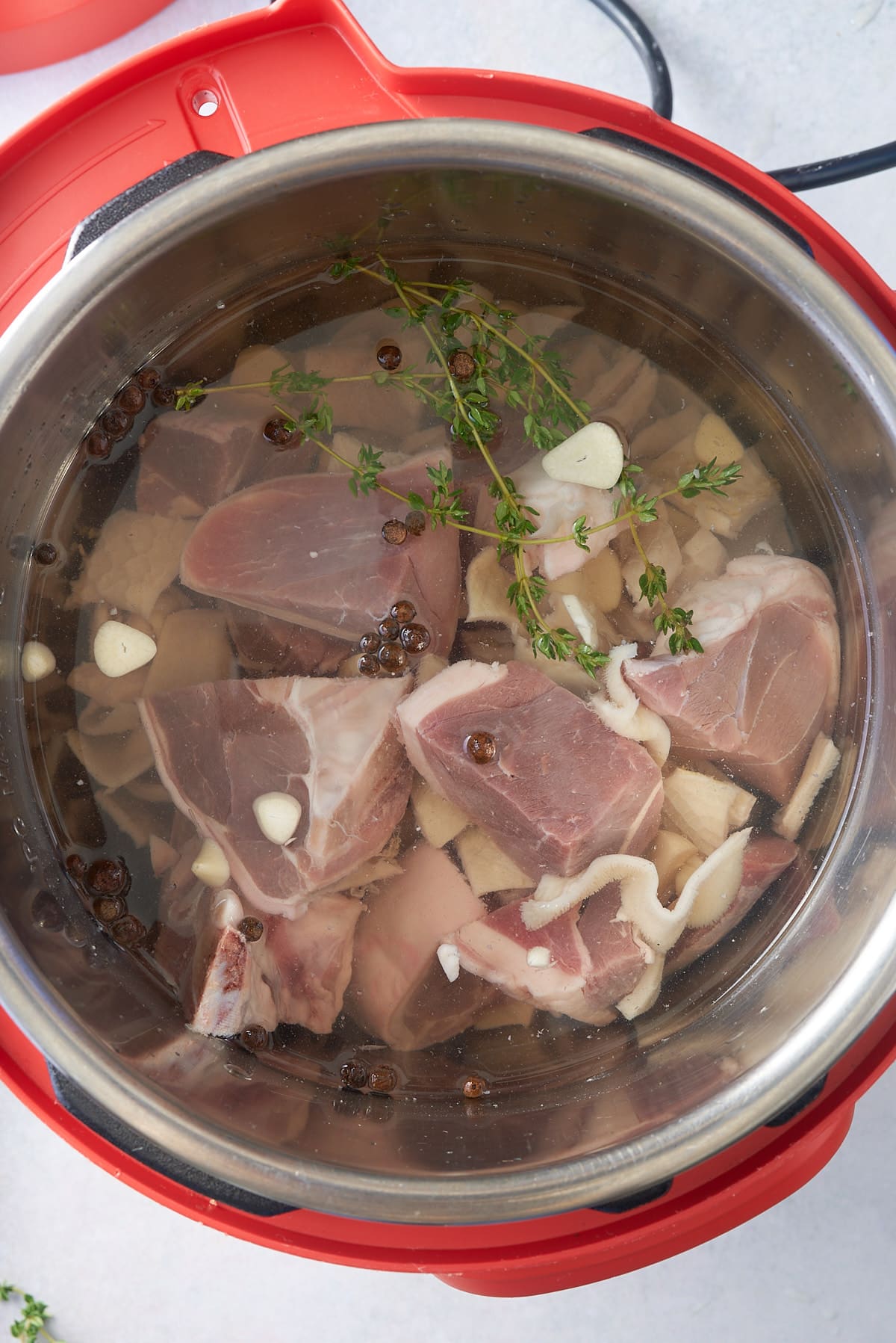 A pressure cooker bowl filled with diced goats meat, tripe, sliced garlic, sprigs of thyme and allspice berries, topped up with water.