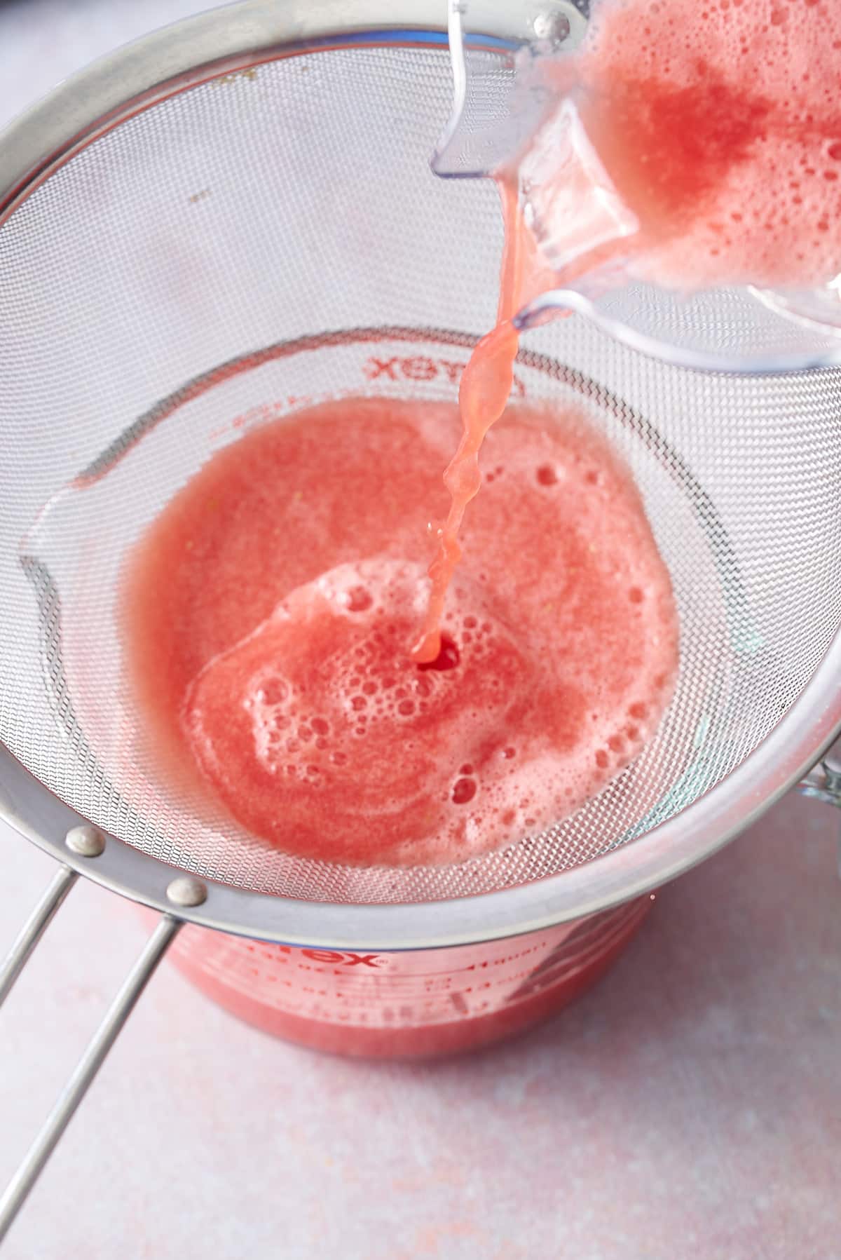 Watermelon juice being poured through a fine sieve into a glass jug.