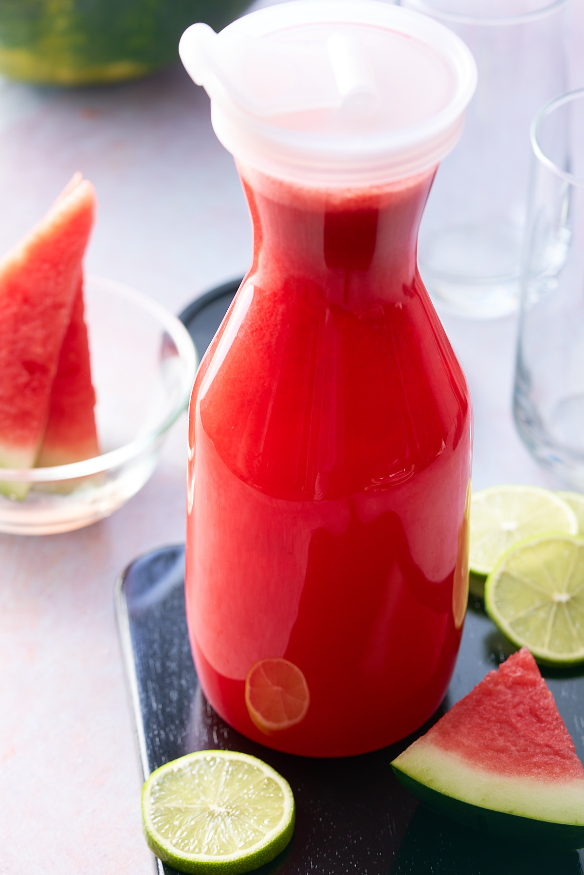 A glass jug filled with watermelon juice with wedges of fresh watermelon and slices of lime set alongside.