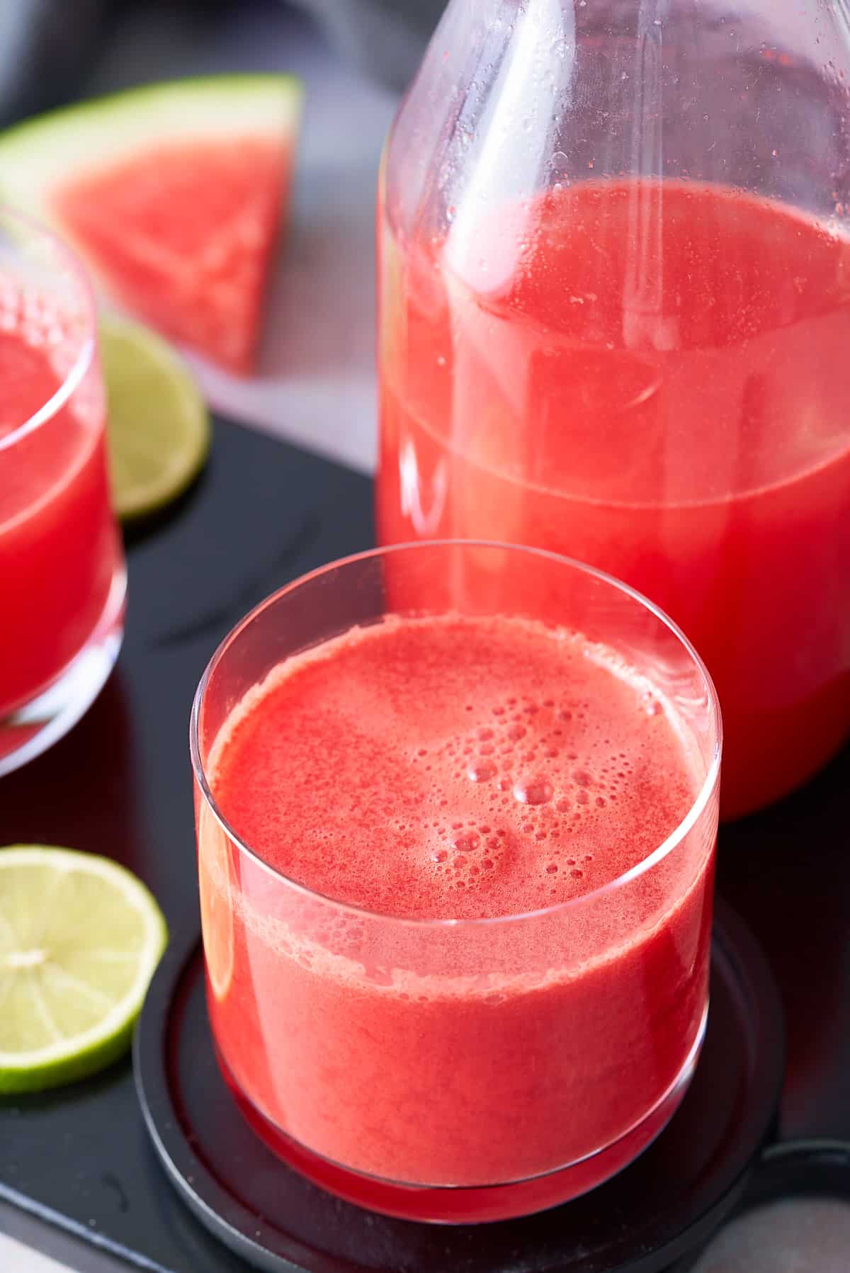 A glass filled with watermelon juice with a jug of watermelon juice, wedges of fresh watermelon and slices of lime set alongside.