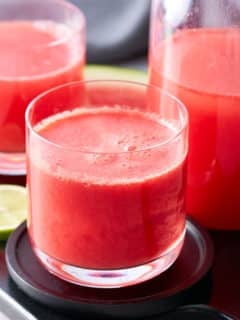 Two glasses filled with watermelon juice with a jug of watermelon juice and slices of lime set alongside.