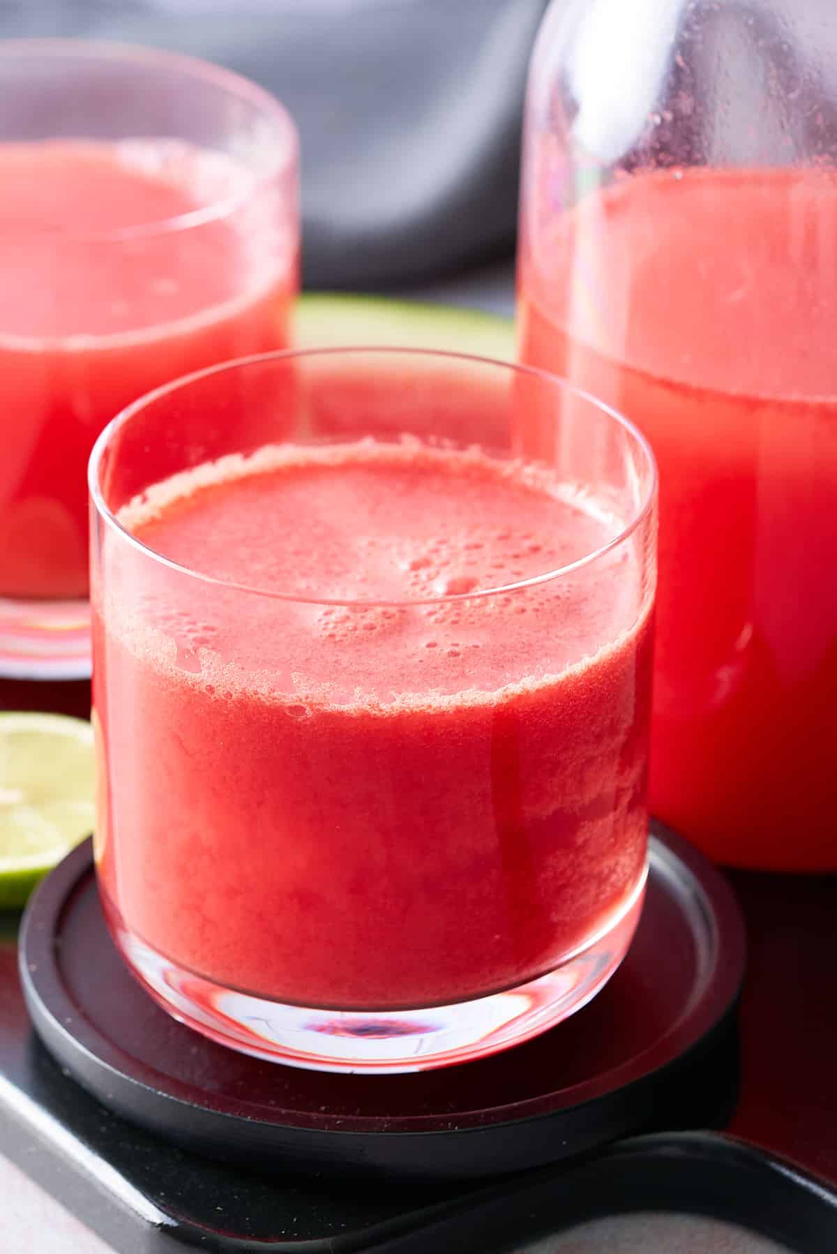 Two glasses filled with watermelon juice with a jug of watermelon juice and slices of lime set alongside.