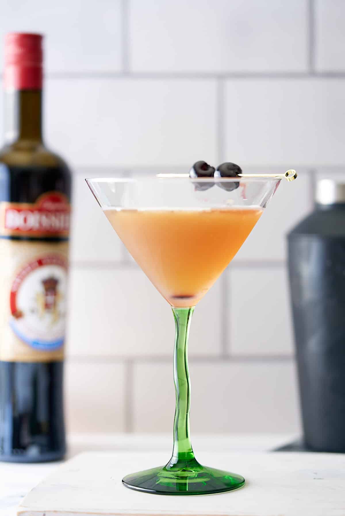 A bourbon martini cocktail in a cocktail glass served with two cocktail cherries, with a bottle of bourbon and cocktail shaker in the background.