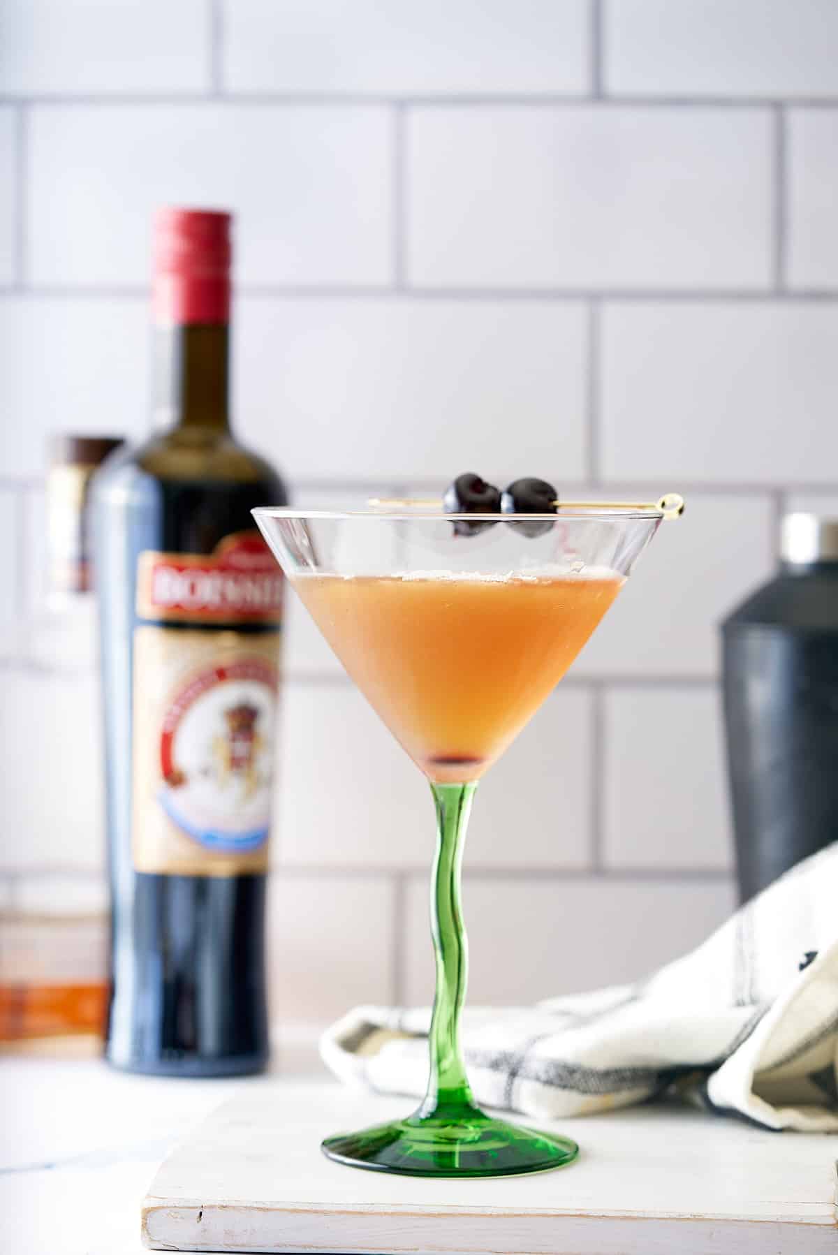 A bourbon martini cocktail in a cocktail glass served with two cocktail cherries, with a bottle of bourbon and cocktail shaker in the background.