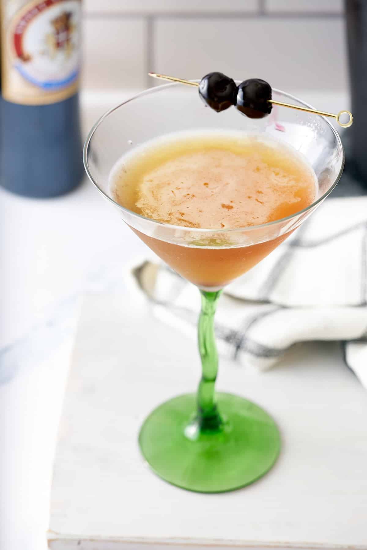 A bourbon martini cocktail in a cocktail glass served with two cocktail cherries, with a bottle of bourbon in the background.