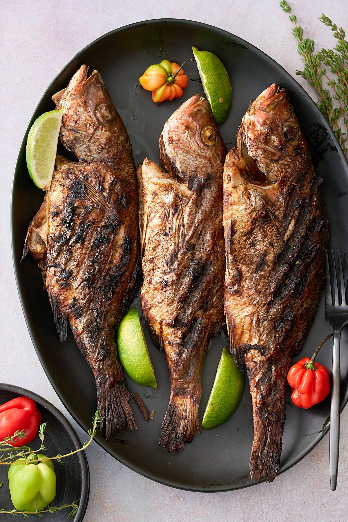 A black serving platter topped with 3 whole snapper fish in a Jamaican jerk marinade garnished with Scotch Bonnet peppers, wedges of lime and sprigs of thyme.