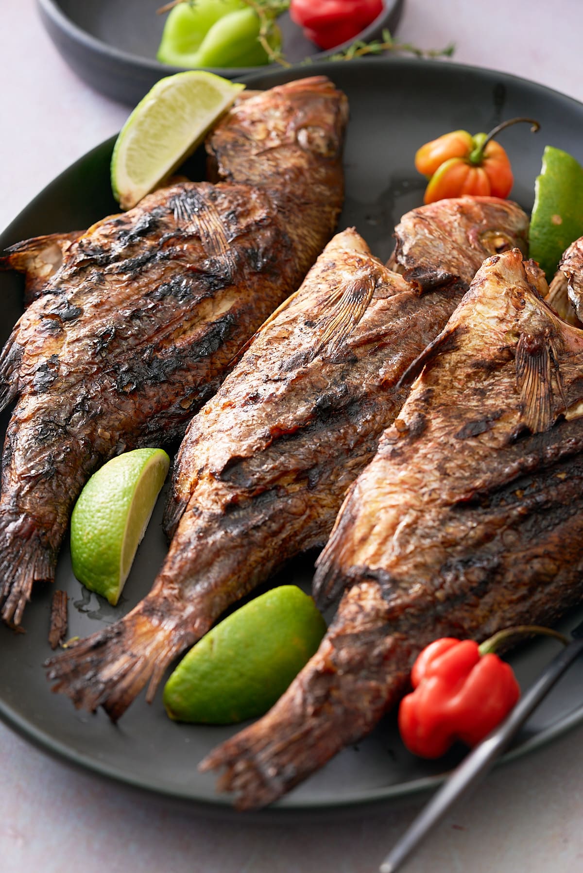 A black serving platter topped with 3 whole snapper fish in a Jamaican jerk marinade garnished with Scotch Bonnet peppers, wedges of lime and sprigs of thyme.