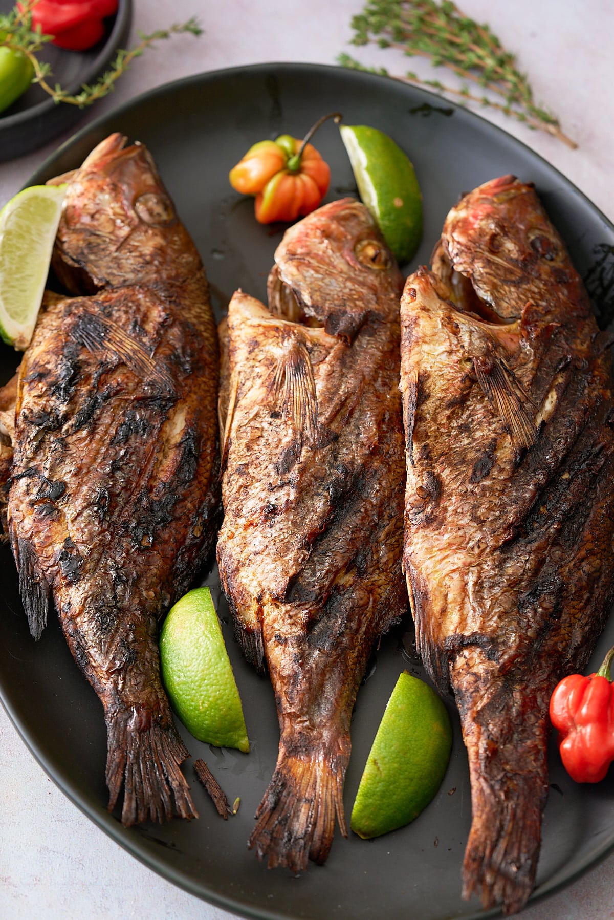 A black serving platter topped with 3 whole snapper fish in a Jamaican jerk marinade garnished with Scotch Bonnet peppers, wedges of lime and sprigs of thyme.