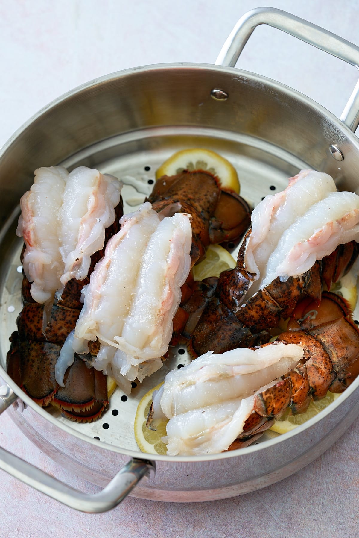 A steamer filled with raw lobster tails on top of fresh slices of lemon.