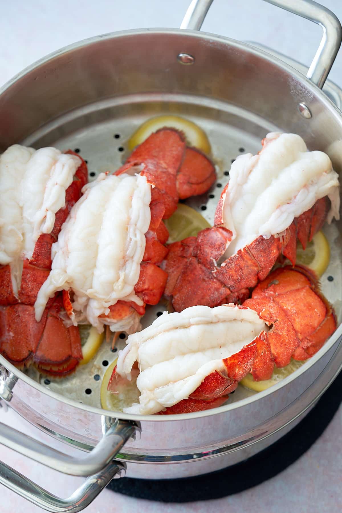 Four cooked lobster tails sitting in a steamer on top of slices of lemon.