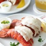 Two white serving plates topped with two steamed lobster tails, served with melted butter, a wedge of lemon and garnished with fresh parsley.