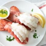 A white serving plate topped with two steamed lobster tails, served with melted butter, a wedge of lemon and garnished with fresh parsley.