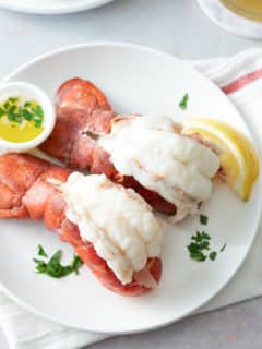 A white serving plate topped with two steamed lobster tails, served with melted butter, a wedge of lemon and garnished with fresh parsley.