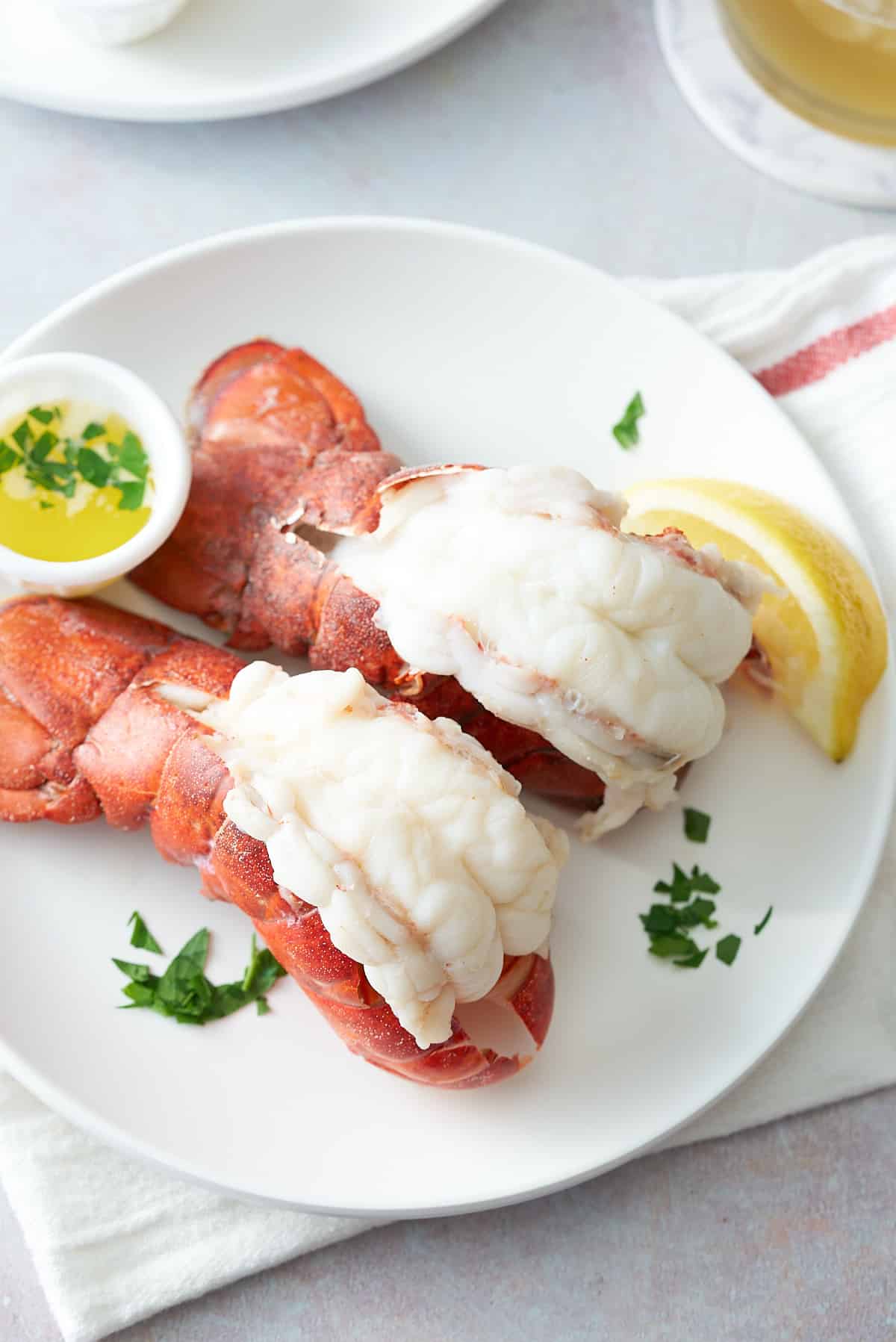 A white serving plate topped with two steamed lobster tails, served with melted butter, a wedge of lemon and garnished with fresh parsley.