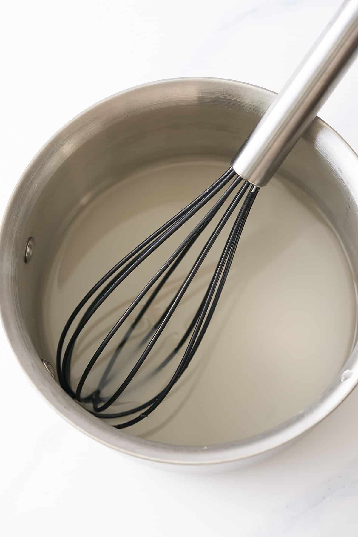 A saucepan filled with water and granulated sugar with a whisk.