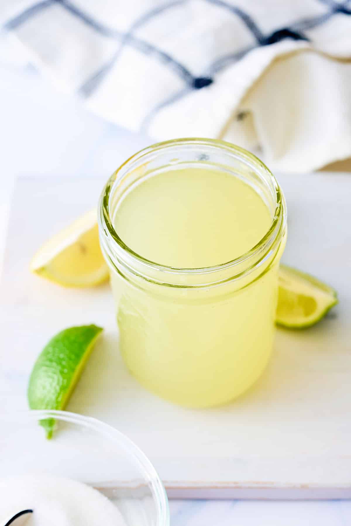 A glass jar filled with sweet and sour mix set on a board with wedges of lemon and lime set alongside.