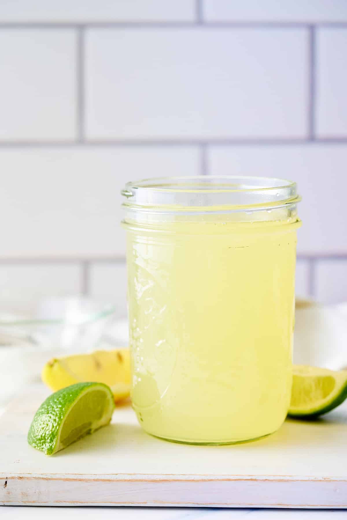 A glass jar filled with sweet and sour mix set on a board with wedges of lemon and lime set alongside.