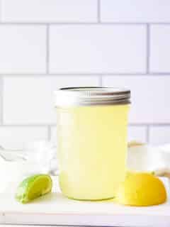 A glass jar filled with sweet and sour mix set on a board with wedges of lemon and lime set alongside.