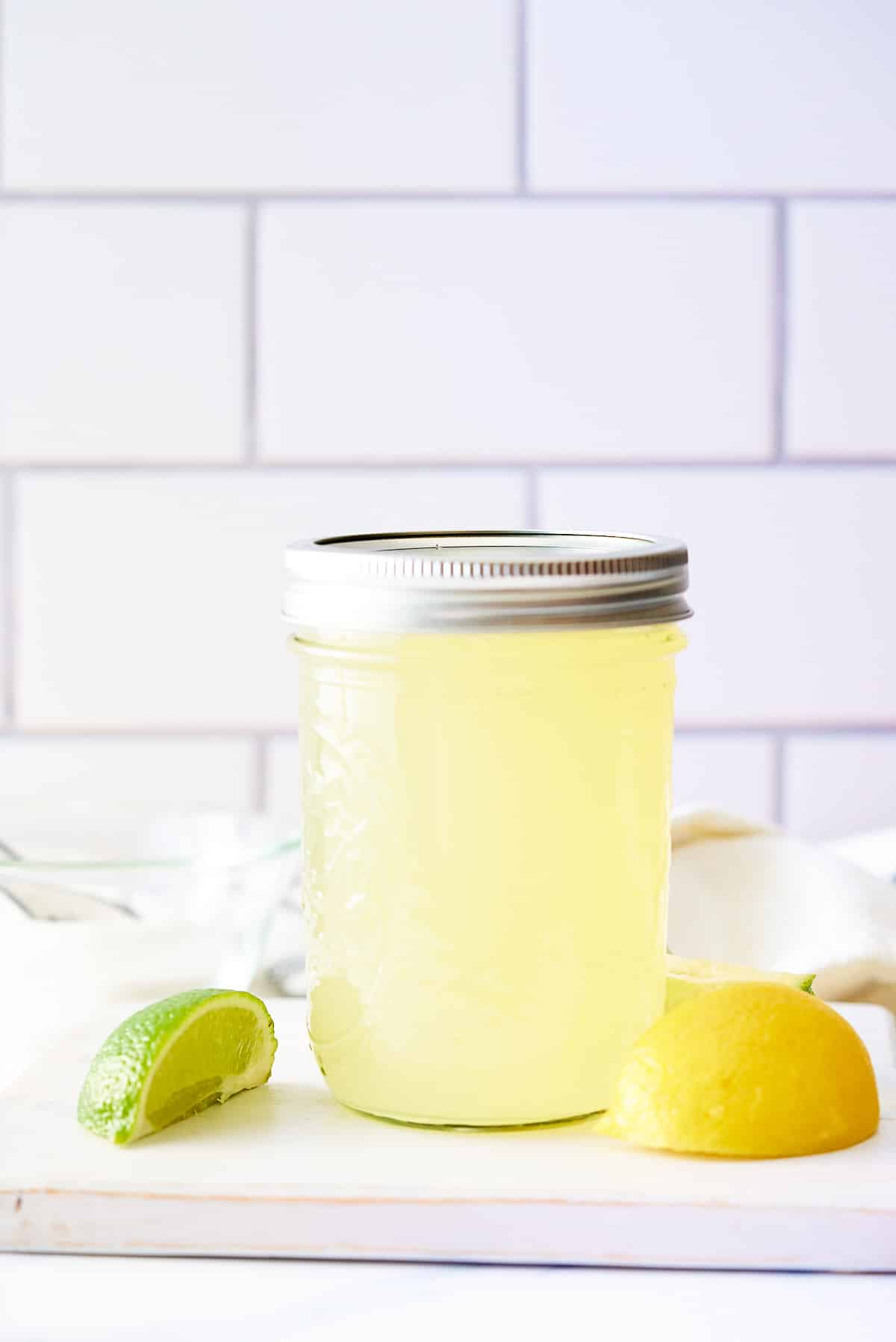 A glass jar filled with sweet and sour mix set on a board with wedges of lemon and lime set alongside.