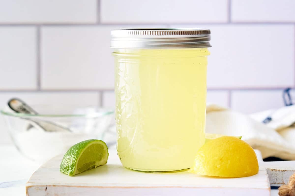 A glass jar filled with sweet and sour mix set on a board with wedges of lemon and lime set alongside.