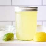 A glass jar filled with sweet and sour mix set on a board with wedges of lemon and lime set alongside.