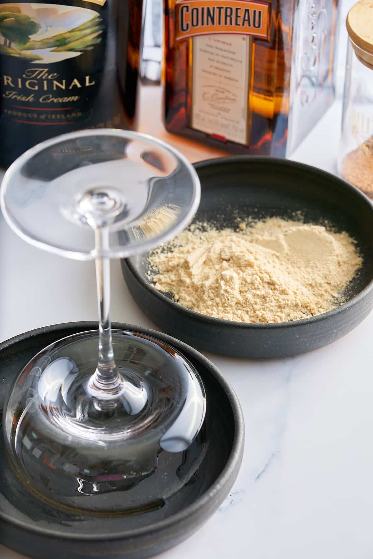 A black dish filled with sugar and Graham cracker crumbs and a second dish filled with whiskey with a glass rim being set into the whiskey.