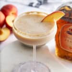 Peach cobbler cocktail in a Graham cracker sugar rimmed glass, garnished with a slice of fresh peach, with a bottle of peach whiskey set alongside.