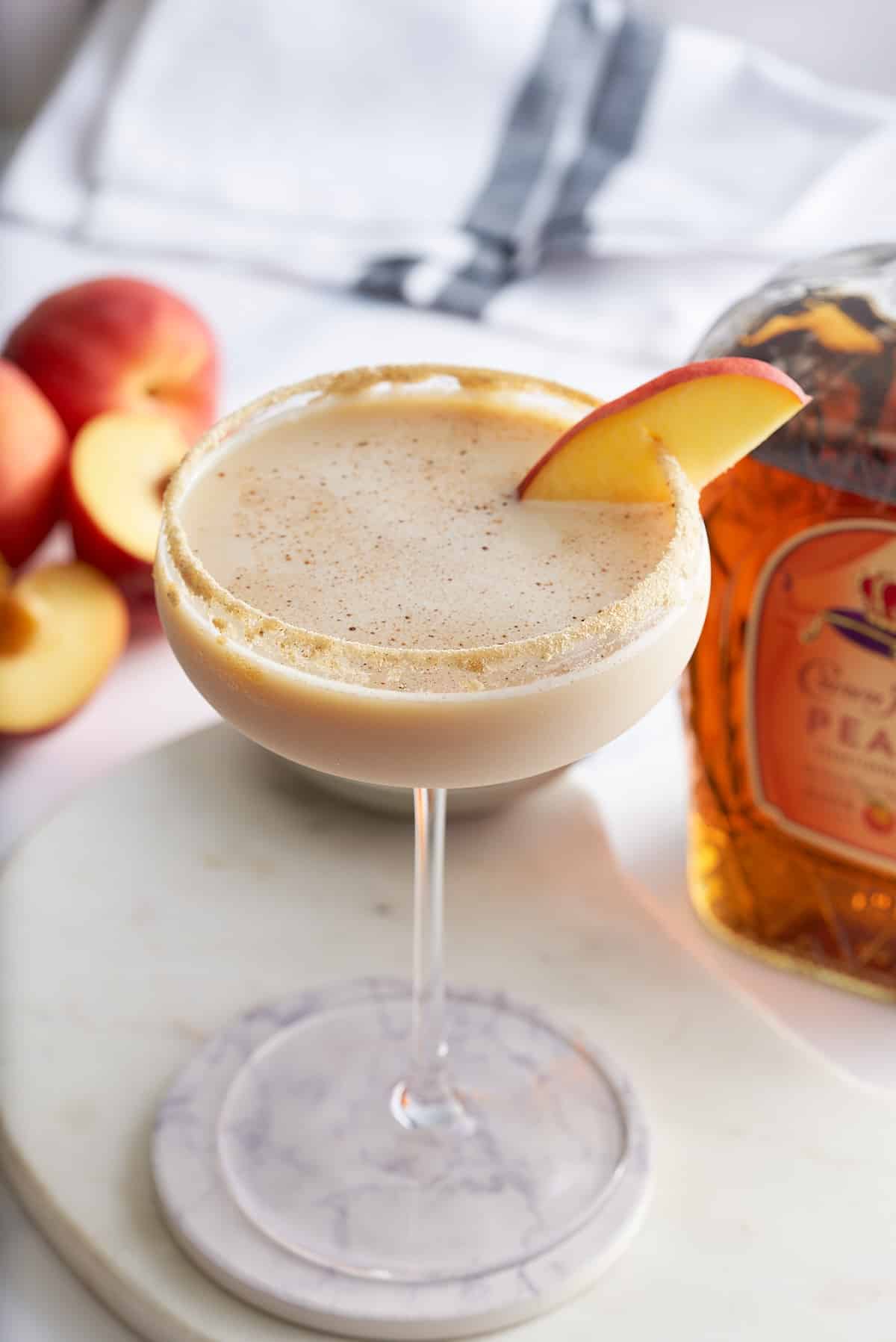 Peach cobbler cocktail in a Graham cracker sugar rimmed glass, garnished with a slice of fresh peach, with a bottle of peach whiskey set alongside.