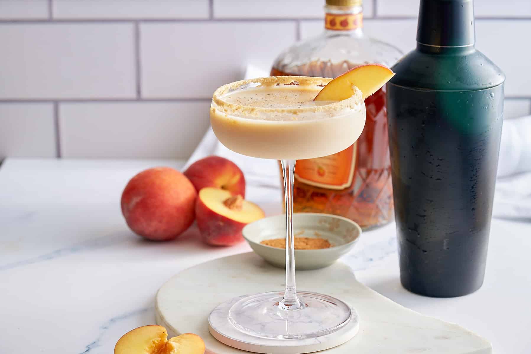 Peach cobbler cocktail in a Graham cracker sugar rimmed glass, garnished with a slice of fresh peach, with a black cocktail shaker, a bottle of peach whiskey and whole peaches set alongside.