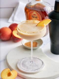 Peach cobbler cocktail in a Graham cracker sugar rimmed glass, garnished with a slice of fresh peach, with a black cocktail shaker, a bottle of peach whiskey and whole peaches set alongside.