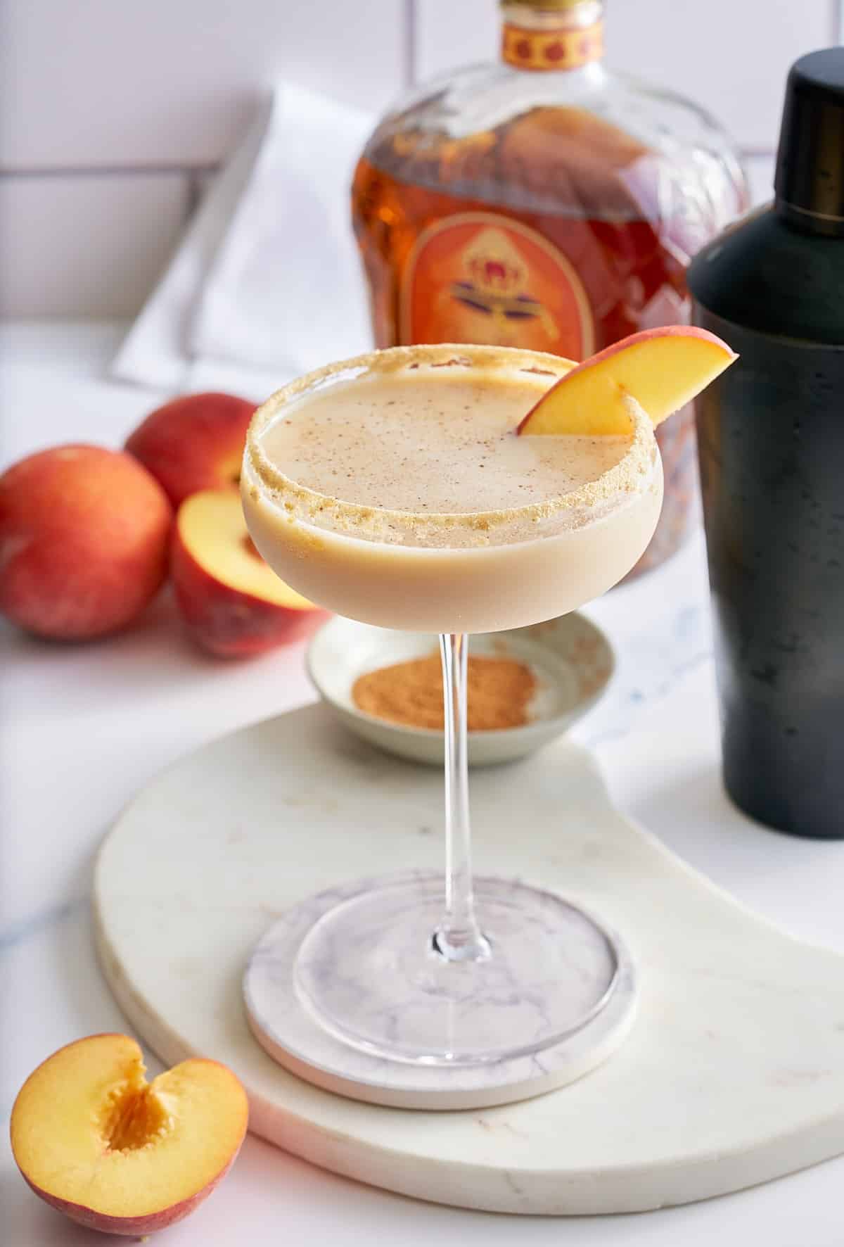 Peach cobbler cocktail in a Graham cracker sugar rimmed glass, garnished with a slice of fresh peach, with a black cocktail shaker, a bottle of peach whiskey and whole peaches set alongside.