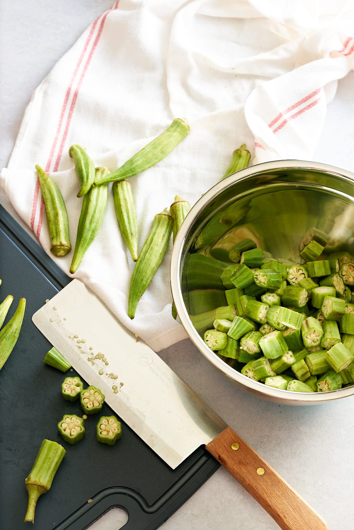 A chopping board with knife and whole and sliced okra, with a silver bowl of sliced okra pieces set alongside.