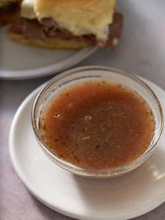 A glass bowl filled with au jus with some roast beef sliders set alongside.