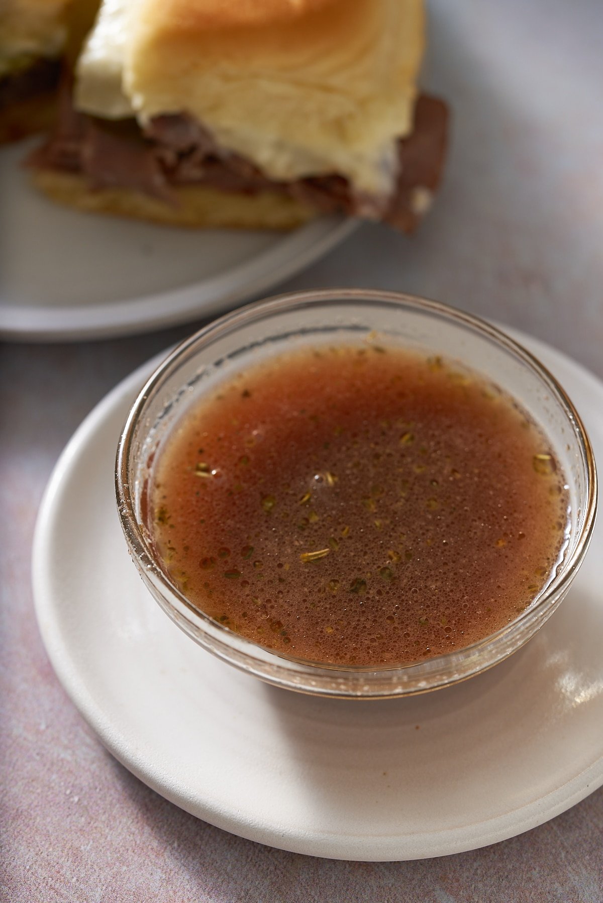 A glass bowl filled with au jus with some roast beef sliders set alongside.