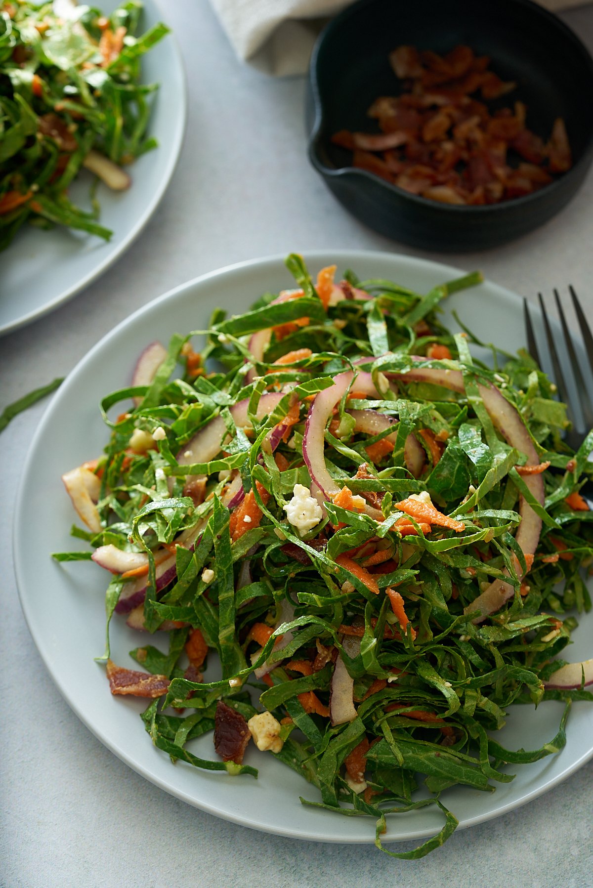 A white plate of fresh collard green salad with a bowl of bacon vinaigrette set alongside.