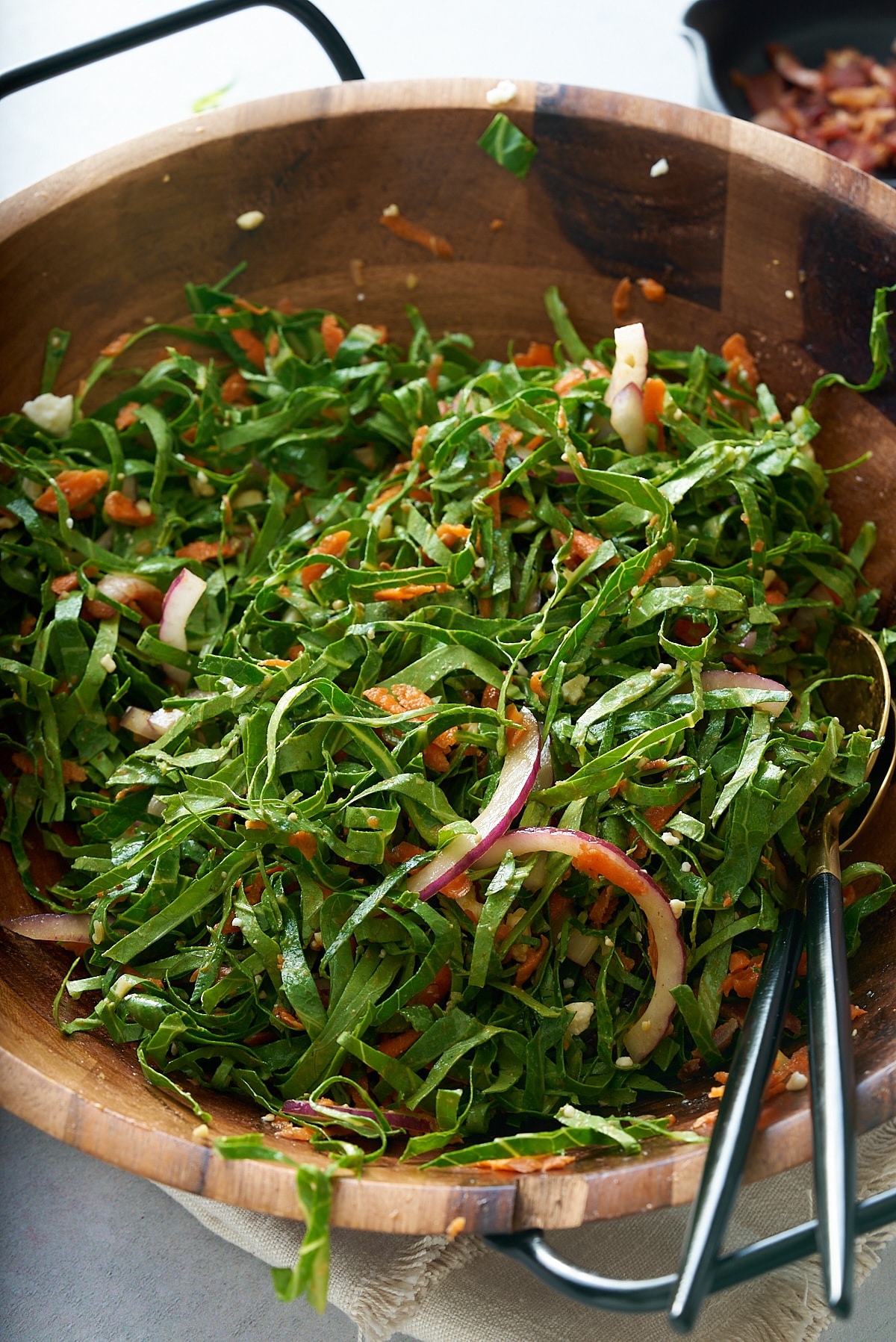 A large wooden salad bowl of fresh collard green salad with salad servers, and a bowl of bacon vinaigrette set alongside.