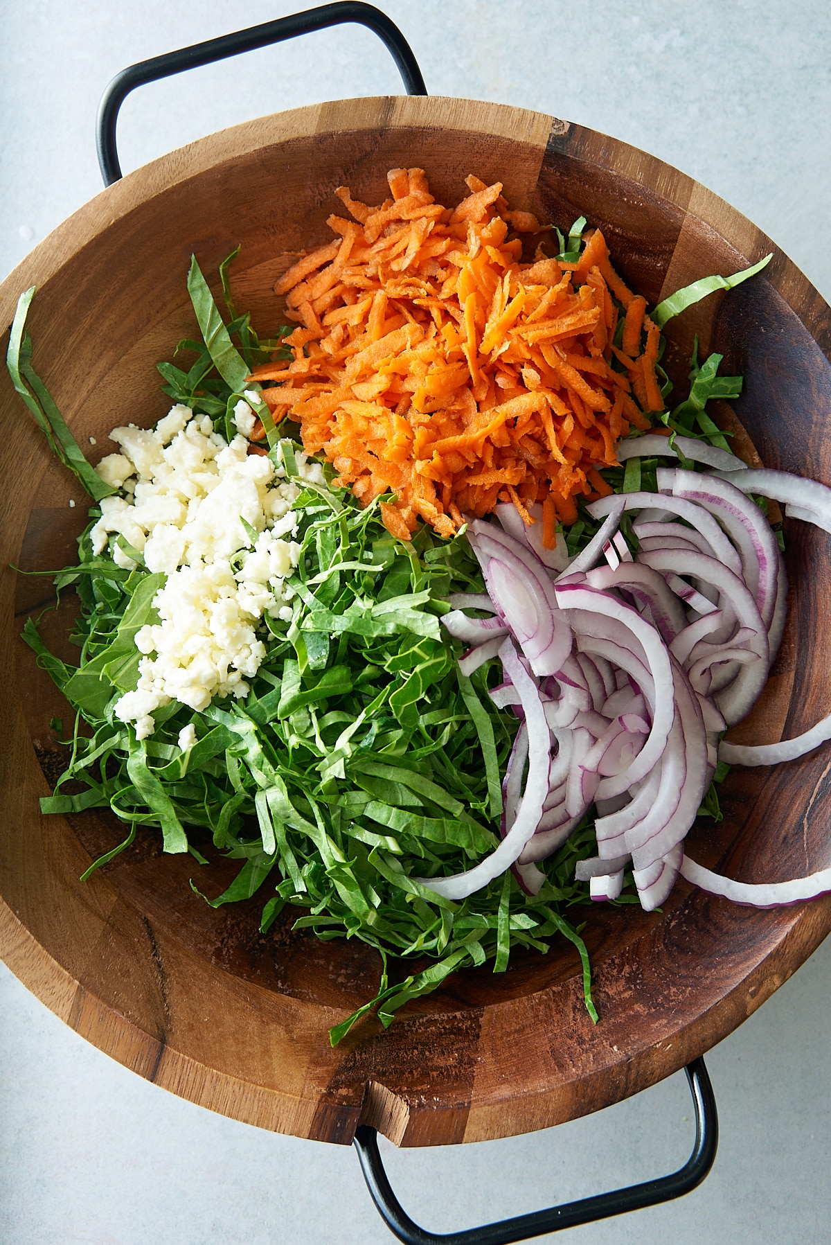A wooden salad bowl filled with sliced salad vegetables, including collard greens, carrots, red onion, and some crumbled feta cheese.