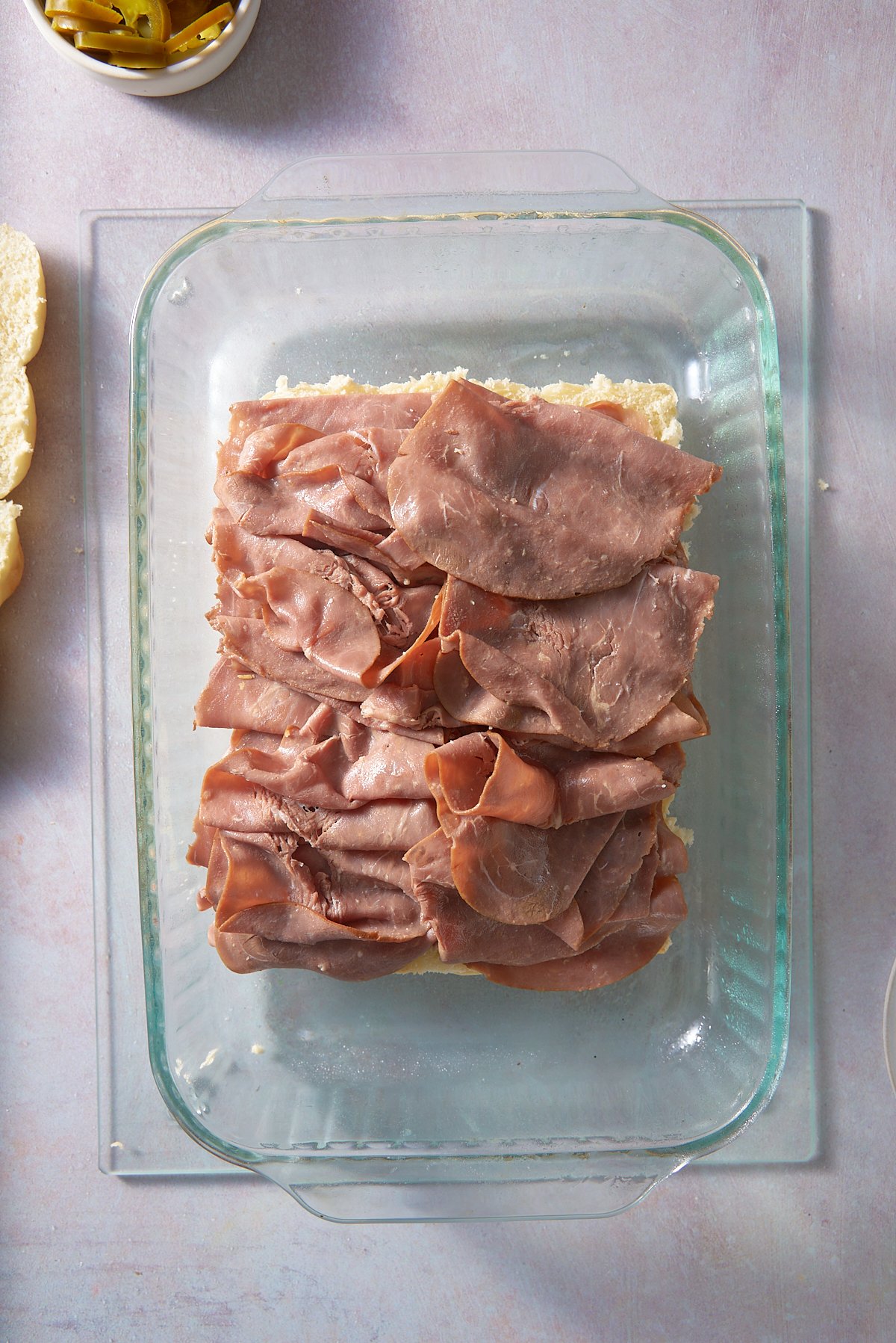 A glass oven proof dish with a the bottom half of 12 slider buns set on the bottom topped with thin slices of roast beef.