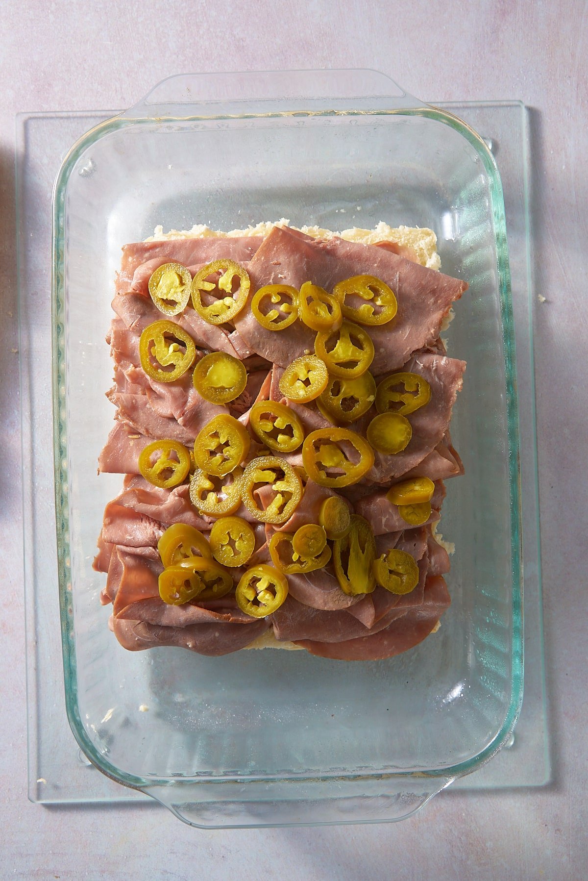 A glass oven proof dish with a the bottom half of 12 slider buns set on the bottom topped with thin slices of roast beef and sliced jalapenos.