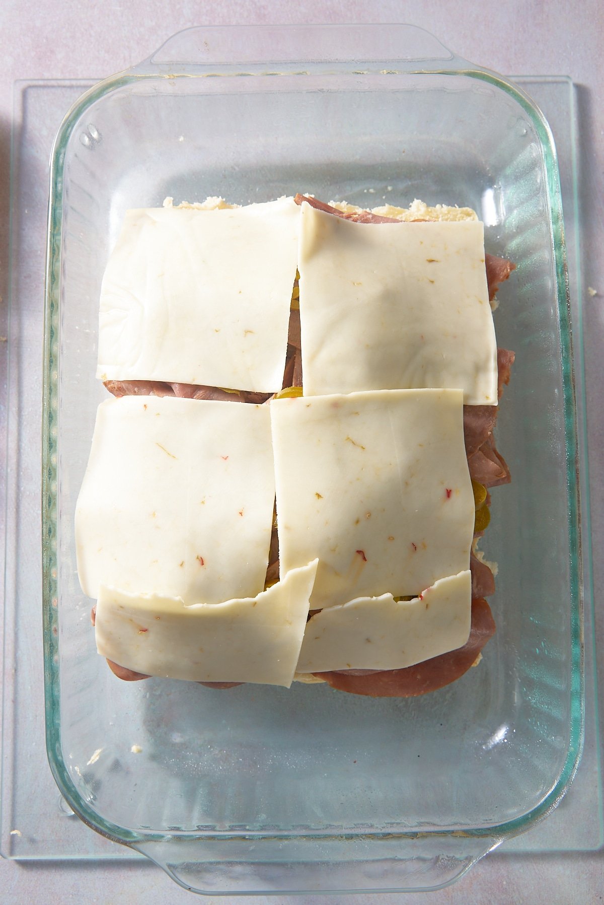 A glass oven proof dish with a the bottom half of 12 slider buns set on the bottom topped with thin slices of roast beef and jalapenos, topped with slices of pepper jack cheese.