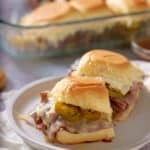 Toasted roast beef sliders on a white plate, with a glass dish of sliders set alongside.