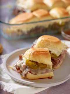 Toasted roast beef sliders on a white plate, with a glass dish of sliders set alongside.