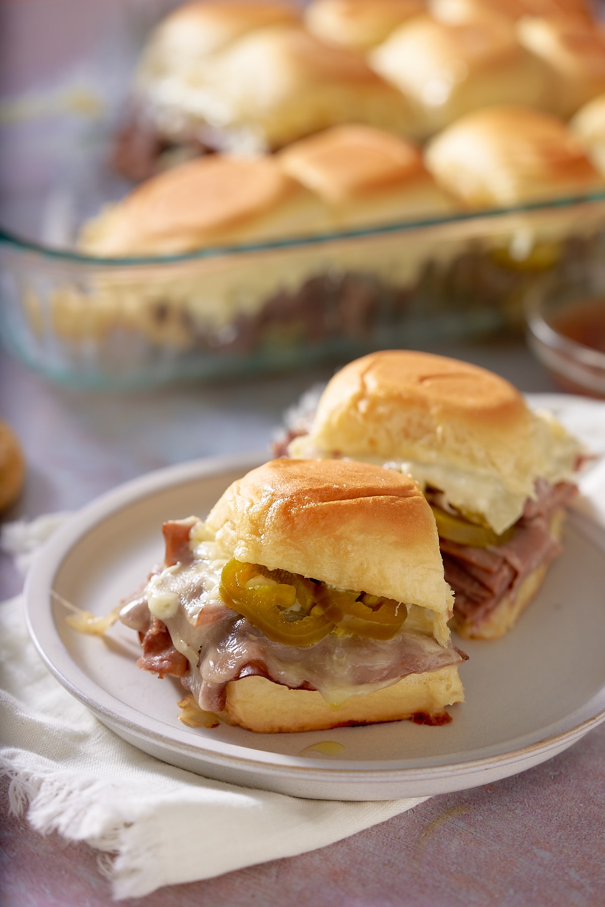 Toasted roast beef sliders on a white plate, with a glass dish of sliders set alongside.