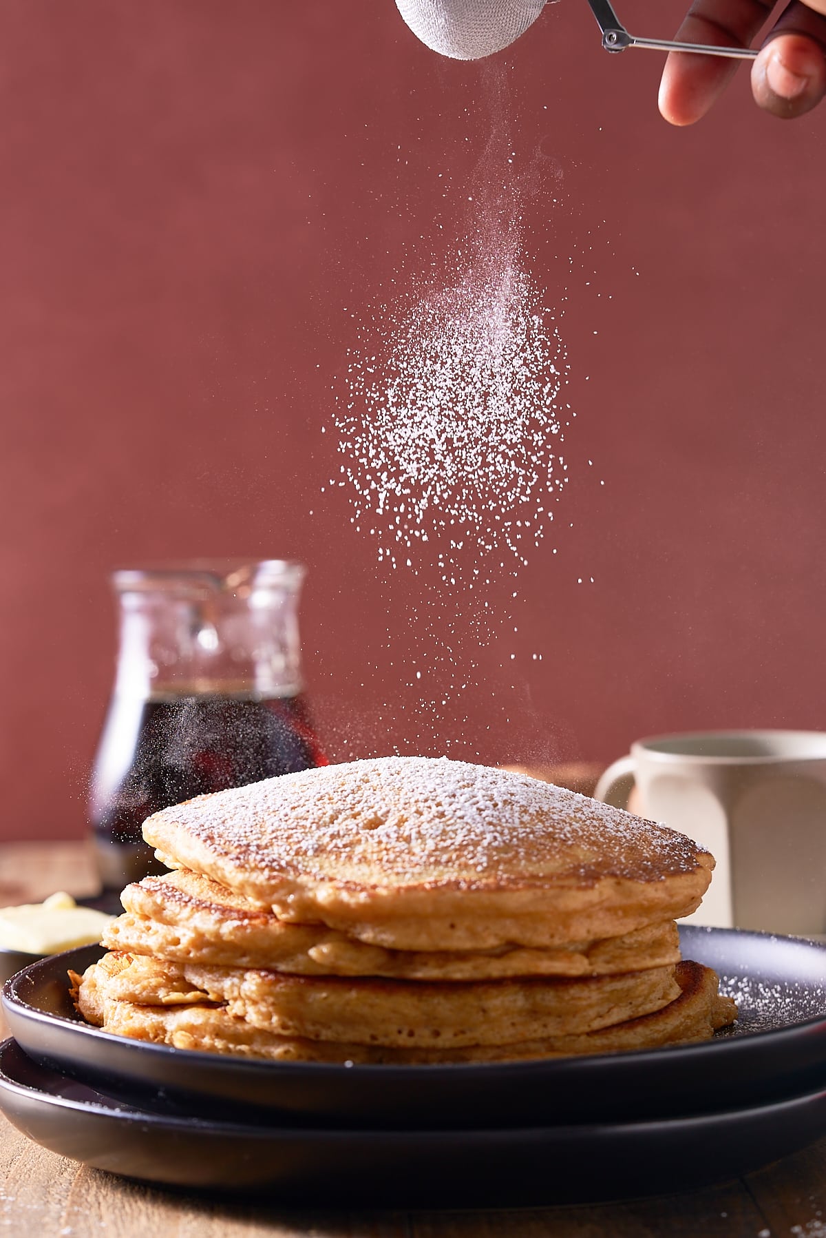 A stack of sweet potato pancakes on a black plate, being dusted with icing sugar, and with a jug of maple syrup served alongside.