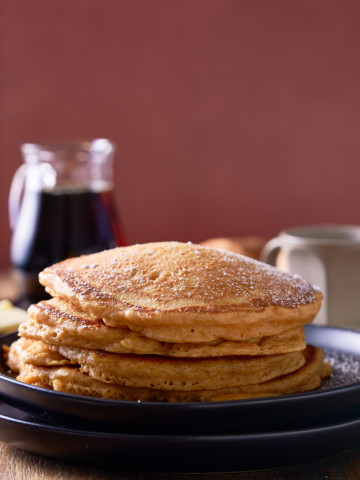A stack of sweet potato pancakes on a black plate with a jug of maple syrup served alongside.