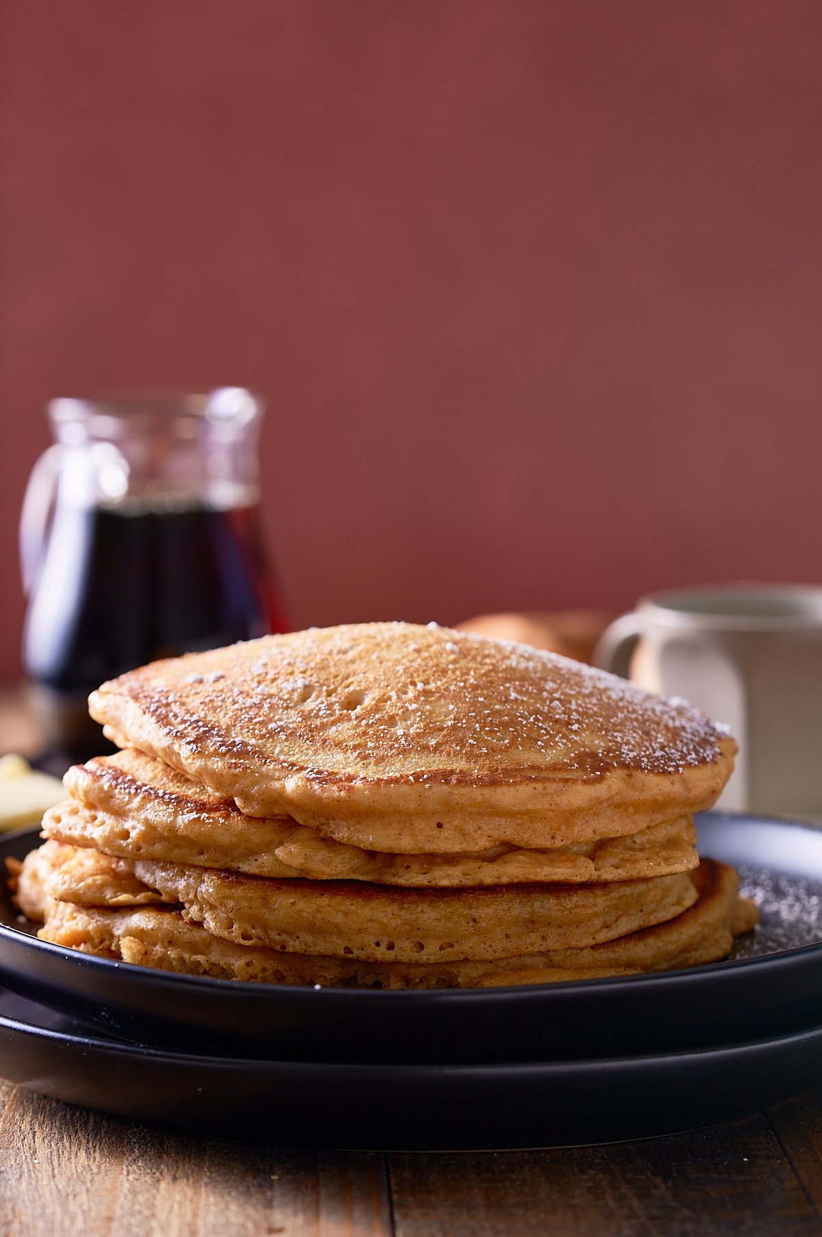A stack of sweet potato pancakes on a black plate with a jug of maple syrup served alongside.